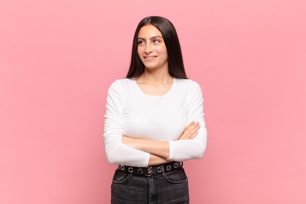 Mujer bonita joven que se siente feliz, orgullosa y esperanzada, preguntándose o pensando, mirando hacia arriba para copiar el espacio con los brazos cruzados