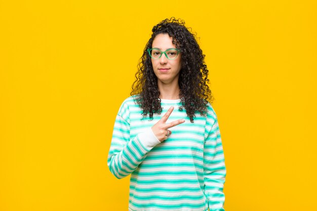 Mujer bonita joven que se siente feliz, positiva y exitosa, con la mano haciendo forma de V sobre el pecho, mostrando la victoria o la paz contra la pared naranja