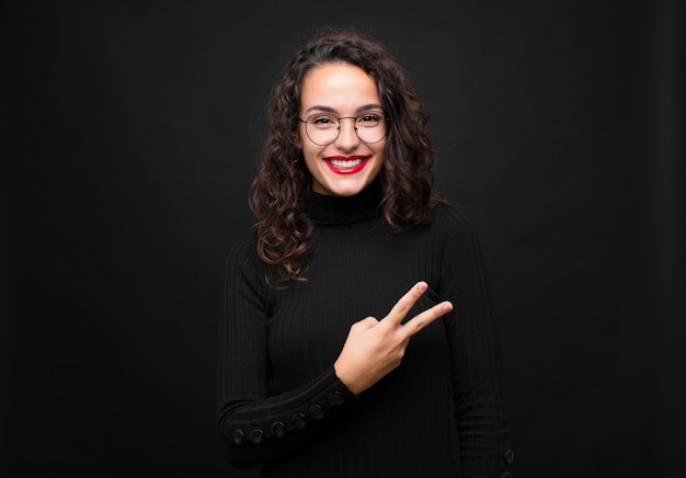 Mujer bonita joven que se siente feliz, positiva y exitosa, con la mano haciendo forma de V sobre el pecho, mostrando la victoria o la paz contra la pared negra.