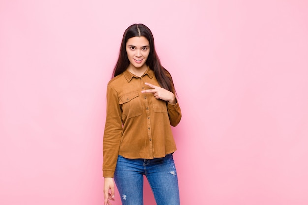 Mujer bonita joven que se siente feliz, positiva y exitosa, con la mano haciendo forma de V sobre el pecho, mostrando victoria o paz sobre la pared rosa