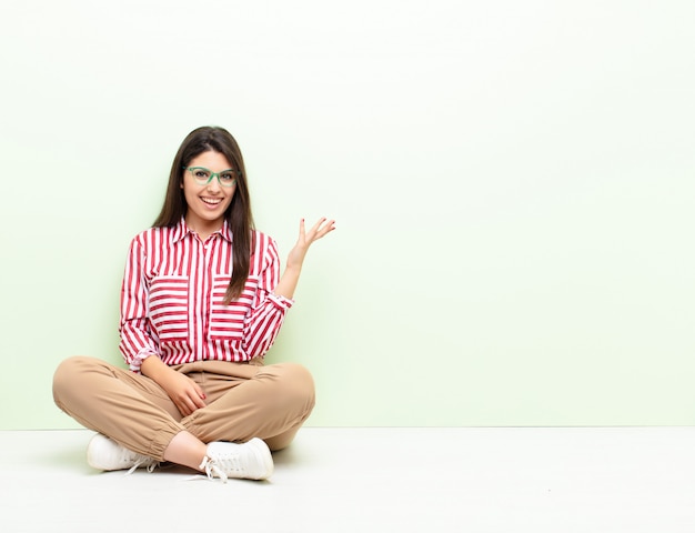 Mujer bonita joven que se siente feliz, sorprendida y alegre, sonriendo con actitud positiva, dándose cuenta de una solución o idea sentada en el suelo