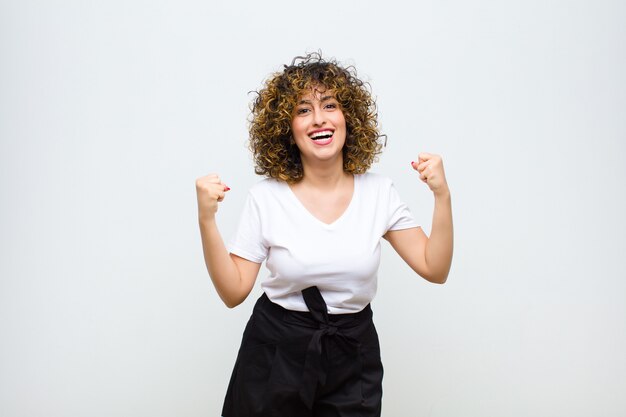 Mujer bonita joven que se siente feliz, sorprendida y orgullosa, gritando y celebrando el éxito con una gran sonrisa contra la pared blanca