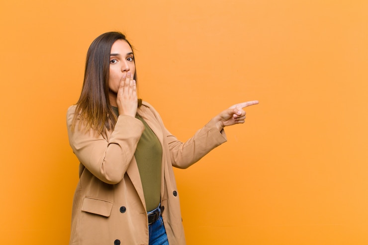 Foto mujer bonita joven que se siente feliz, sorprendida y sorprendida, cubriendo la boca con la mano y apuntando al espacio de copia lateral aislado sobre la pared naranja
