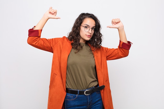 Foto mujer bonita joven que se siente orgullosa, arrogante y confiada, luciendo satisfecha y exitosa, apuntando a sí misma contra la pared blanca