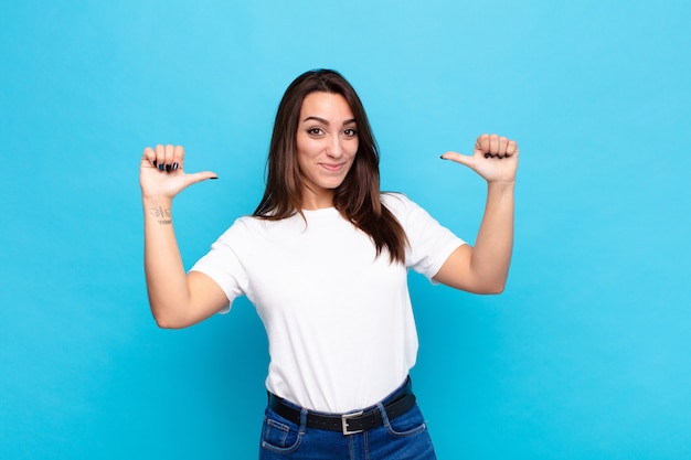 Mujer bonita joven que se siente orgullosa, arrogante y segura, que parece satisfecha y exitosa, apuntando a sí misma contra la pared azul