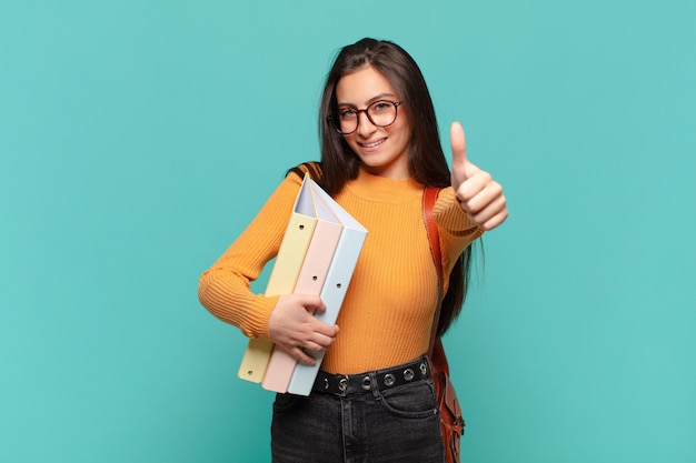 Mujer bonita joven que se siente orgullosa, despreocupada, segura y feliz, sonriendo positivamente con los pulgares hacia arriba. concepto de estudiante