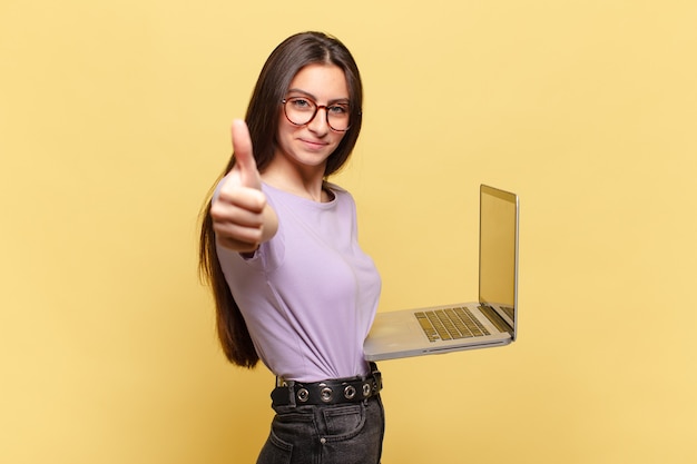 Mujer bonita joven que se siente orgullosa, despreocupada, segura y feliz, sonriendo positivamente con los pulgares hacia arriba. concepto de laptop