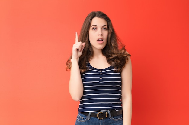 Mujer bonita joven que se siente orgullosa y sorprendida, apuntando a sí misma con confianza, sintiéndose como la número uno exitosa contra la pared roja