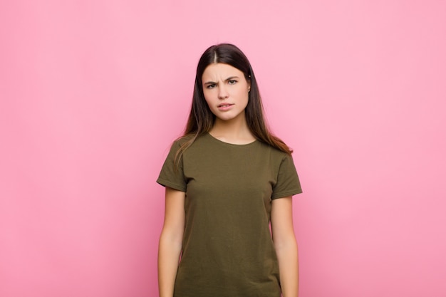 Mujer bonita joven que se siente perpleja y confundida, con una expresión muda y aturdida mirando algo inesperado contra la pared rosa