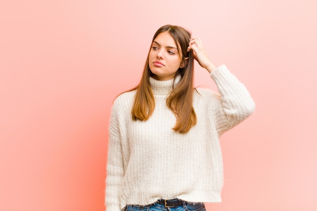 Mujer bonita joven que se siente perpleja y confundida, rascándose la cabeza y mirando a un lado contra la pared rosa