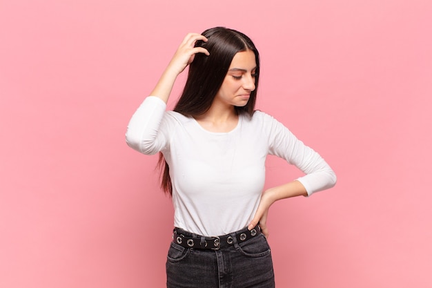 Mujer bonita joven que se siente perpleja y confundida, rascándose la cabeza y mirando hacia un lado