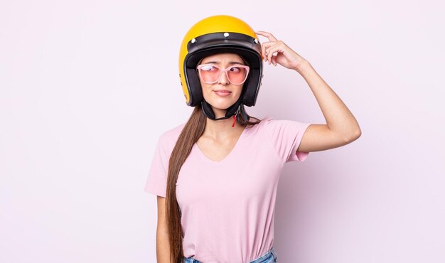 Mujer bonita joven que se siente perpleja y confundida, rascándose la cabeza. motociclista y casco