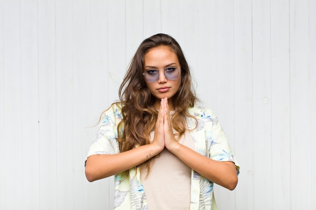 Mujer bonita joven que se siente preocupada, esperanzada y religiosa, orando fielmente con las palmas presionadas, pidiendo perdón