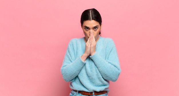 Mujer bonita joven que se siente preocupada, esperanzada y religiosa, orando fielmente con las palmas presionadas, pidiendo perdón