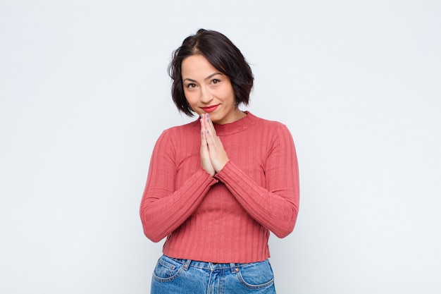 Mujer bonita joven que se siente preocupada, esperanzada y religiosa, rezando fielmente con las palmas presionadas, pidiendo perdón contra la pared blanca