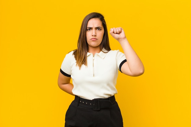 Mujer bonita joven que se siente seria, fuerte y rebelde, levantando el puño, protestando o luchando por la revolución aislada contra la pared naranja