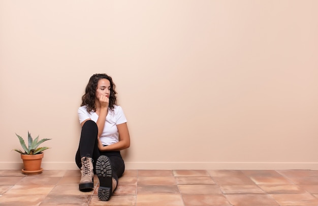 Foto mujer bonita joven que se siente seria, pensativa y preocupada, mirando hacia los lados con la mano presionada contra la barbilla sentada en el piso de una terraza