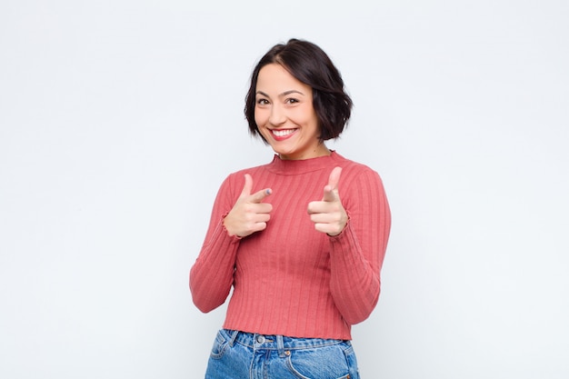 Mujer bonita joven que sonríe con una actitud positiva, acertada, feliz que señala a la cámara, haciendo la muestra del arma con las manos sobre la pared blanca