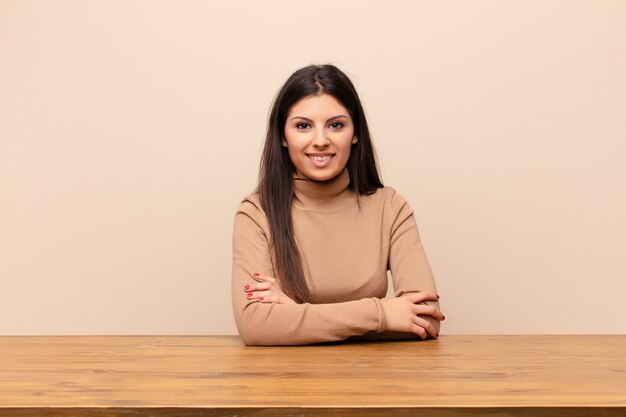 Mujer bonita joven que sonríe con los brazos cruzados y una expresión feliz, confiada y satisfecha, vista lateral sentado en una mesa