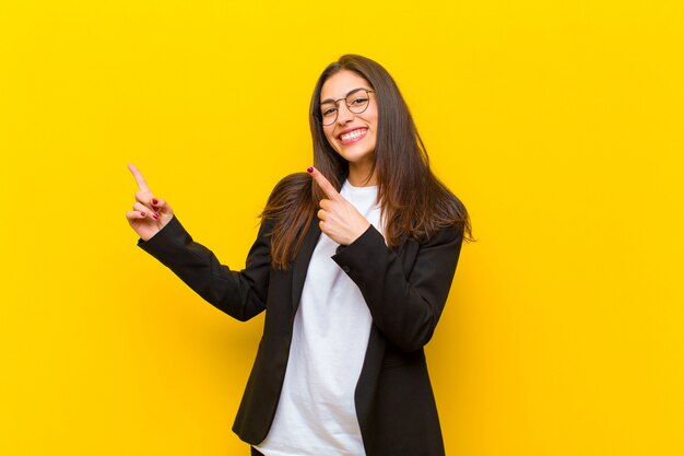 Mujer bonita joven que sonríe feliz y que señala al lado y hacia arriba con ambas manos que muestran el objeto en espacio de la copia contra la pared anaranjada