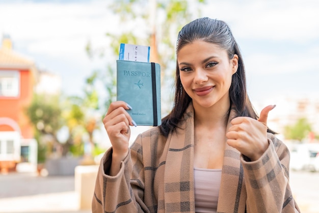 Foto mujer bonita joven que sostiene un pasaporte al aire libre con los pulgares hacia arriba porque ha sucedido algo bueno