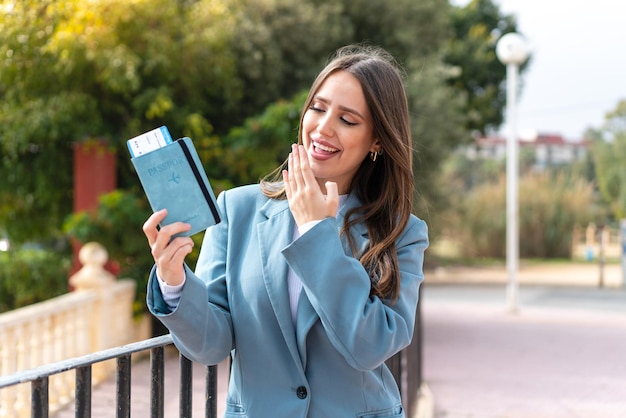 Mujer bonita joven que sostiene un pasaporte al aire libre con sorpresa y expresión facial conmocionada