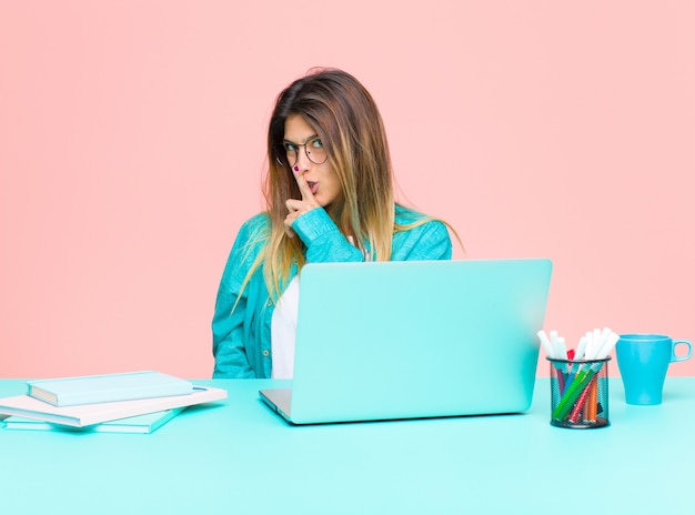 Mujer bonita joven que trabaja con una computadora portátil pidiendo silencio y silencio, gesticulando con el dedo frente a la boca, diciendo shh o guardando un secreto