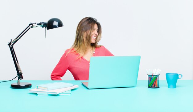 Mujer bonita joven que trabaja con una computadora portátil que parece feliz, alegre y confiada, sonriendo con orgullo y mirando al lado con ambas manos en las caderas