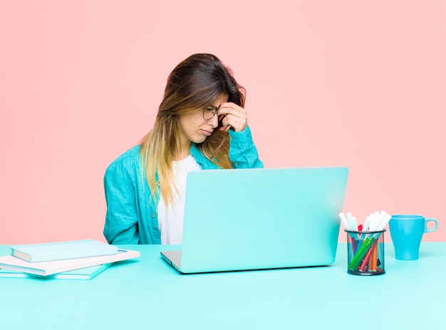 Mujer bonita joven que trabaja con una computadora portátil sintiéndose estresada, infeliz y frustrada, tocando la frente y sufriendo migraña de dolor de cabeza intenso