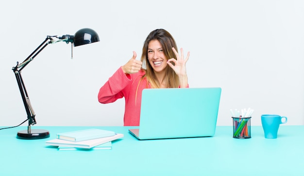 Mujer bonita joven que trabaja con una computadora portátil sintiéndose feliz, asombrada, satisfecha y sorprendida, mostrando gestos bien y pulgares arriba, sonriendo