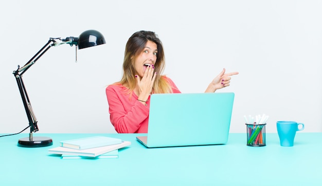 Mujer bonita joven que trabaja con una computadora portátil sintiéndose feliz, sorprendida y sorprendida, cubriendo la boca con la mano y apuntando al espacio de copia lateral