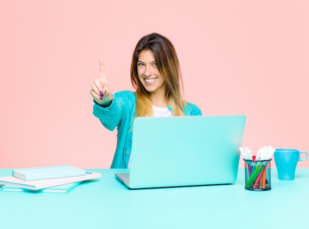 Mujer bonita joven que trabaja con una computadora portátil sonriendo y mirando amigable, mostrando el número uno o primero con la mano hacia adelante, cuenta atrás