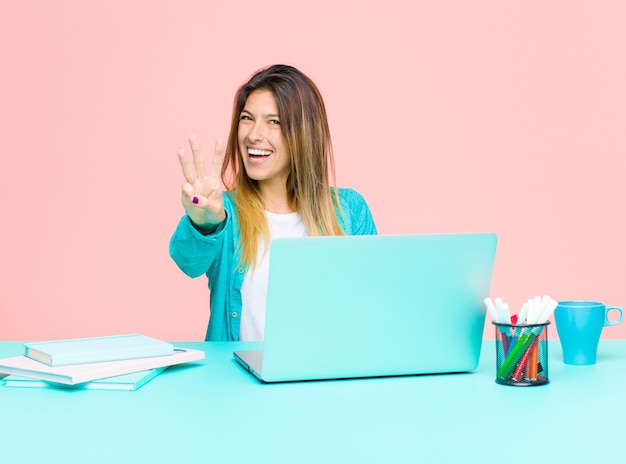 Mujer bonita joven que trabaja con una computadora portátil sonriendo y mirando amigable, mostrando el número tres con la mano hacia adelante, cuenta atrás