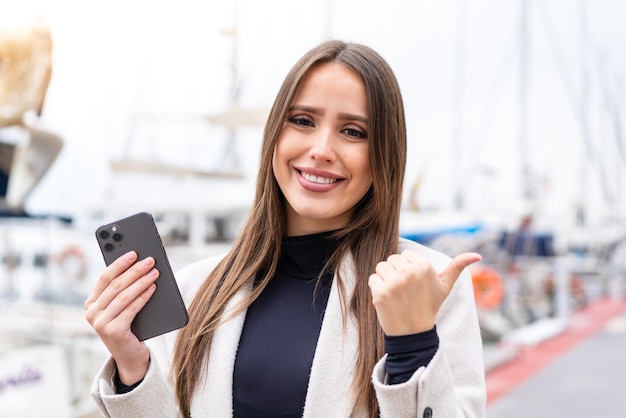 Mujer bonita joven que usa el teléfono móvil al aire libre apuntando hacia un lado para presentar un producto