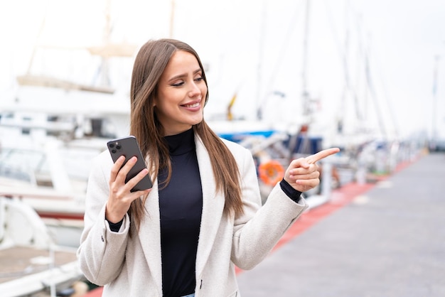 Mujer bonita joven que usa el teléfono móvil al aire libre apuntando hacia un lado para presentar un producto