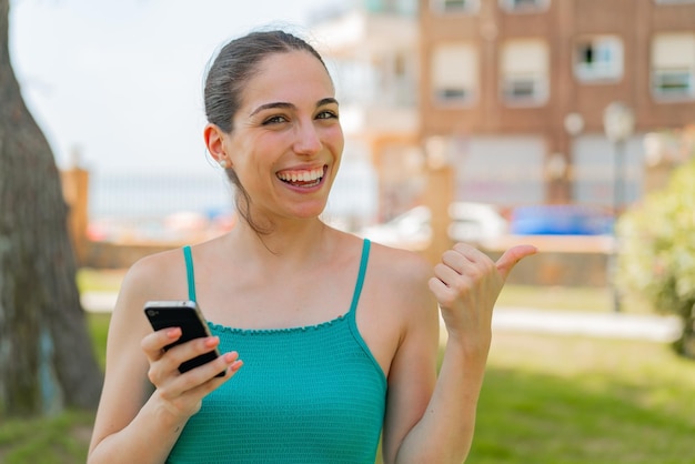 Mujer bonita joven que usa un teléfono móvil al aire libre apuntando hacia un lado para presentar un producto