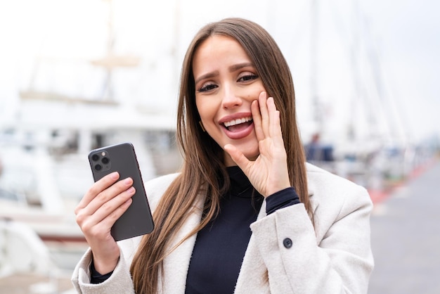 Mujer bonita joven que usa el teléfono móvil al aire libre gritando con la boca abierta