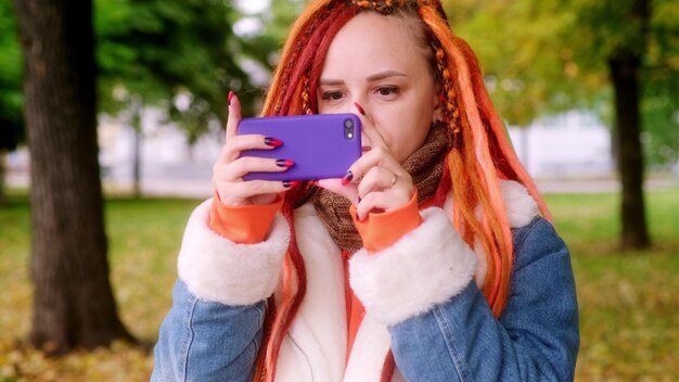 Mujer bonita joven con rastas fotografiando en el teléfono móvil de pie en un parque Turista femenina con colorido peinado multicolor tomando fotos en el gadget