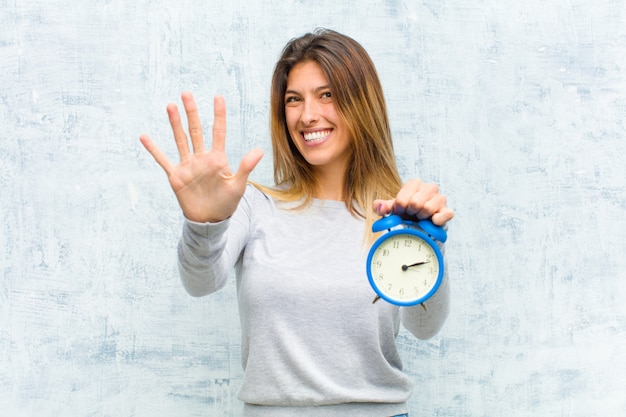 Mujer bonita joven con un reloj de alarma contra la pared del grunge