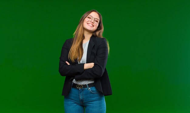 Mujer bonita joven riendo felizmente con los brazos cruzados, con una pose relajada, positiva y satisfecha contra la pared verde