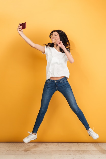 Mujer bonita joven saltando hacer selfie por teléfono con gesto de paz.