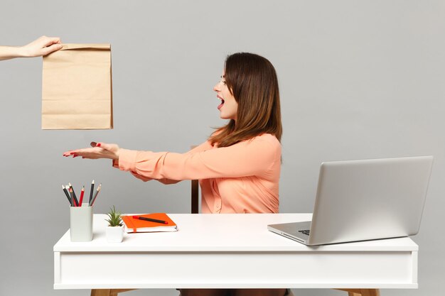 Mujer bonita joven señalando con las manos en una bolsa de papel artesanal marrón clara y vacía para llevar, siéntese a trabajar en el escritorio con una computadora portátil aislada en un fondo gris. Concepto de estilo de vida de carrera empresarial de logro.