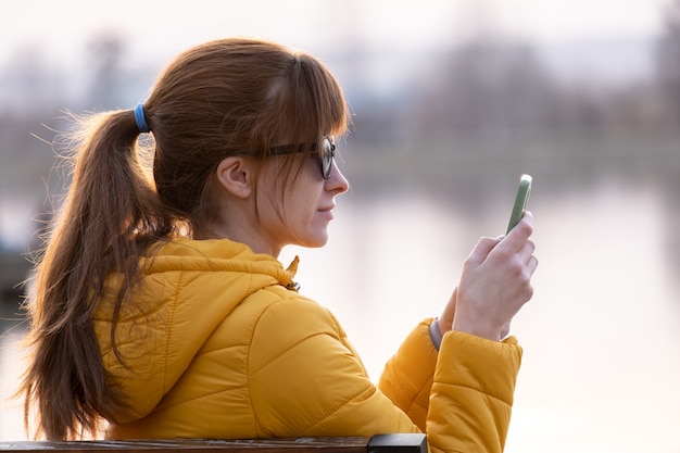 Mujer bonita joven sentada en un banco del parque mirando su sellphone al aire libre por la noche.