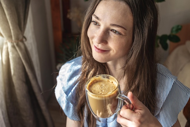 Mujer bonita joven sentada cerca de la ventana y disfrutando de un café fragante por la mañana Concepto de un pasatiempo agradable buen humor y felicidad en cosas pequeñas