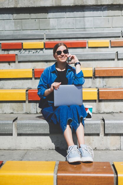Mujer bonita joven sentada con una computadora portátil en la calle del parque moderno usando un teléfono inteligente en el trabajo de TI remoto estilo de verano sonriendo casualmente con gafas