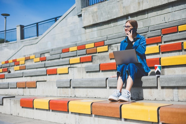 Mujer bonita joven sentada con una computadora portátil en la calle del parque moderno usando un teléfono inteligente en el trabajo de TI remoto estilo de verano sonriendo casualmente con gafas