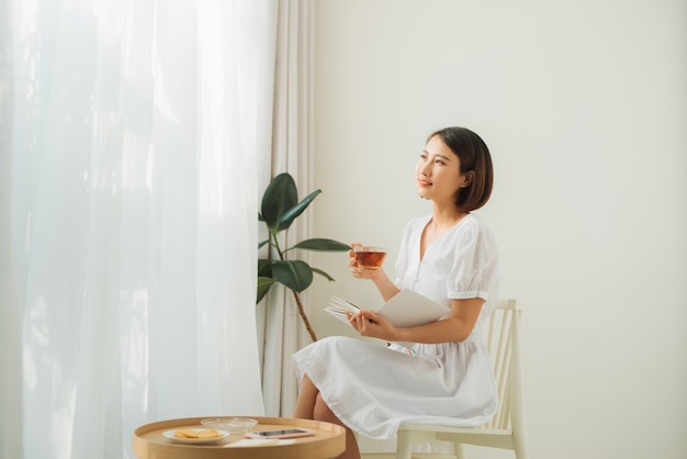 Mujer bonita joven sentada junto a la ventana bebiendo té y leyendo un libro disfruta de descanso