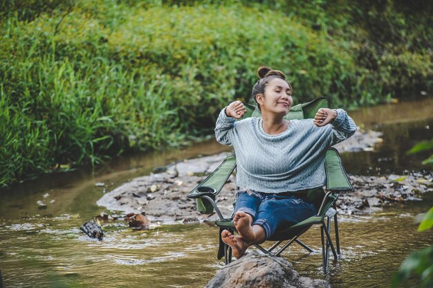 Mujer bonita joven sentada en una silla de camping en el arroyo para relajarse, sonríe en el bosque natural mientras acampa con espacio de copia de felicidad