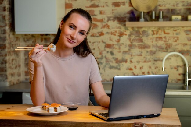 Mujer bonita joven sentada en su cocina comer sushi en la mesa en casa trabajando con una computadora portátil