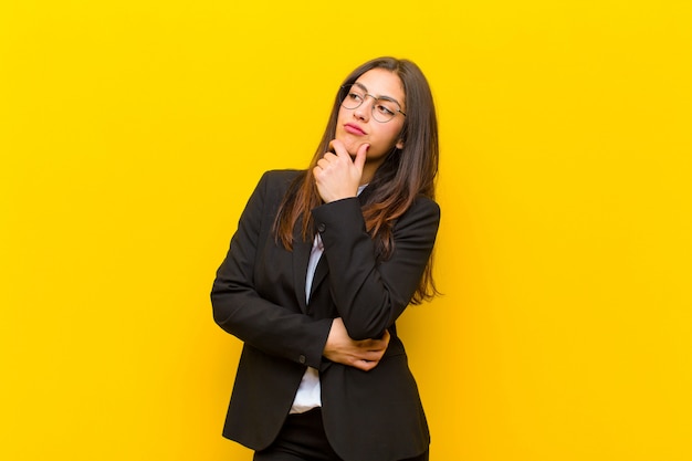 Mujer bonita joven sintiéndose pensativa, preguntándose o imaginando ideas, soñando despierta y mirando a copyspace contra la pared naranja